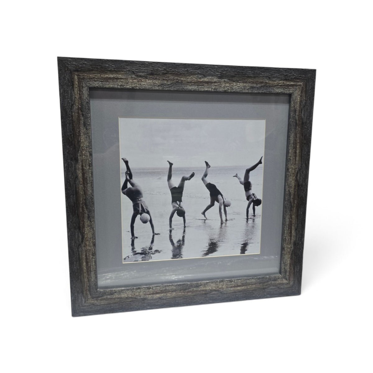 Framed black and white beach photograph showing four people doing handstands along the ocean shoreline