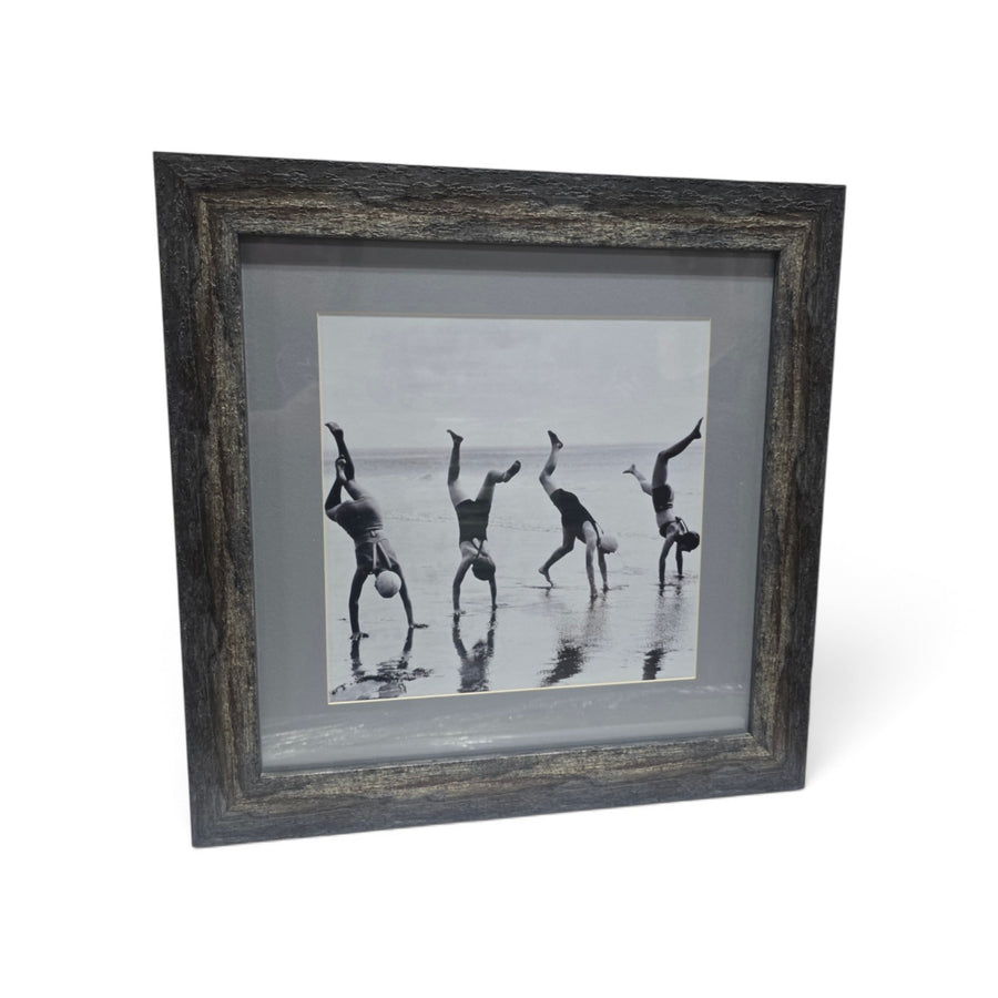 Framed black and white beach photograph showing four people doing handstands along the ocean shoreline